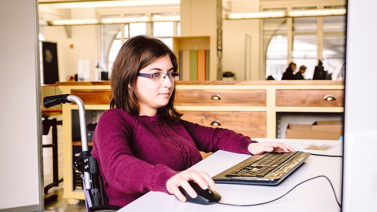 Femme assise dans un fauteuil roulant et travaillant sur un ordinateur. | © Gesellschaftsbilder