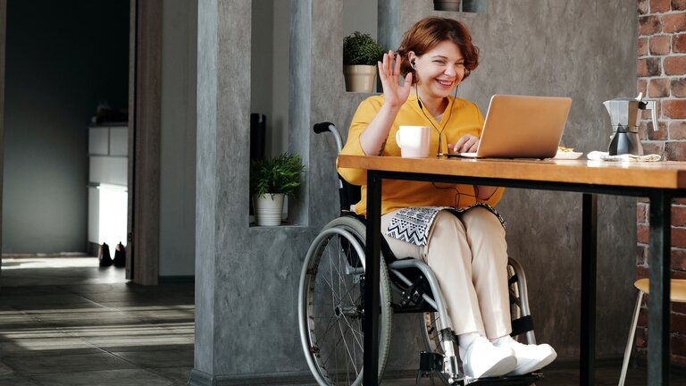 Eine Frau im Rollstuhl winkt in die Kamera ihres Laptops. | © Pexels / Marcus Aurelius