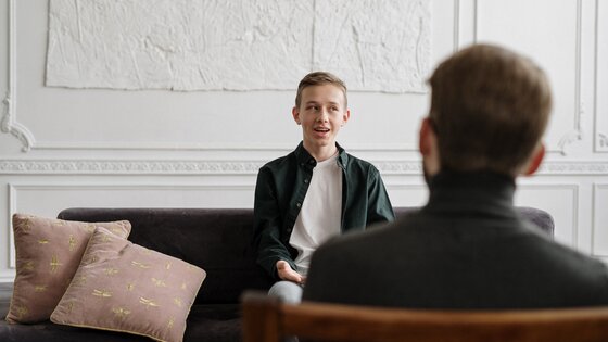 Ein junger Mann mit blondem Haar sitzt auf einem Sofa, ihm gegenüber ein ältere Mann mit Notizblock, der ihn coacht. | © Pexels /Cottonbro