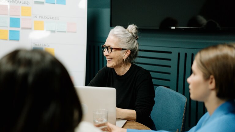 Eine ältere Frau beteiligt sich lächelnd an einer Gesprächsrunde in einem Sitzungszimmer. | © pexels