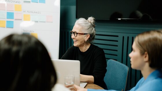 Eine ältere Frau beteiligt sich lächelnd an einer Gesprächsrunde in einem Sitzungszimmer. | © Pexels / Cottonbro