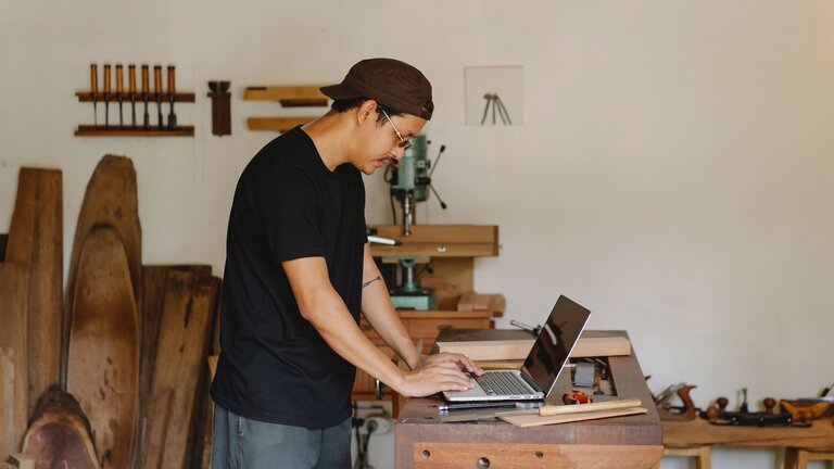 Dans un atelier, un homme travaille sur son ordinateur portable posé sur un établi. | © pexels