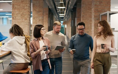 Vier Kolleg:innen sprechen gehend miteinander. Es scheint als ob sie gerade aus einem Meeting kommen. Sie halten Laptop sowie Kaffetassen in den Händen. | © pexels