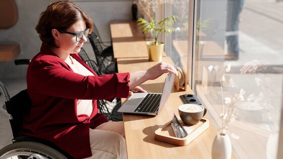 Eine brillentragende Frau im Rollstuhl sitzt in einem Café vor ihrem Laptop.