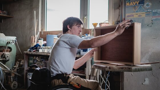 Mann sitzt im Rollstuhl in einer Werkstatt und schleift ein Holzmöbel.