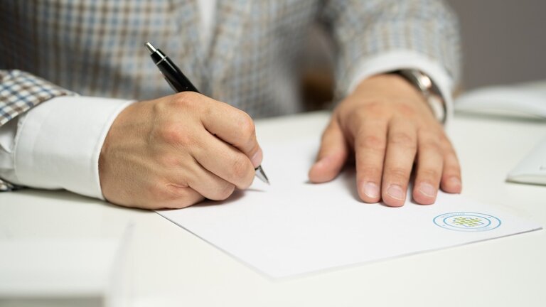 Das Foto zeigt die Hände eines Mannes, der an einem weißen Tisch sitzt und mit einem schwarzen Kugelschreiber ein weißes Blatt Papier unterzeichnet. | © pixabay