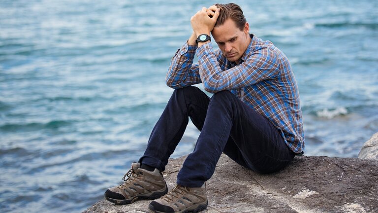 Ein Mann sitzt alleine auf einem Felsen vor einem See oder dem Meer. Er stützt den Kopf auf seine Hände und sieht sehr unglücklich aus. | © pexels