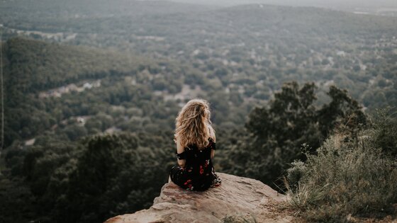 Eine Frau sitzt alleine in der Natur auf einem Aussichtspunkt und schaut runter in den Wald.  | © Unsplash