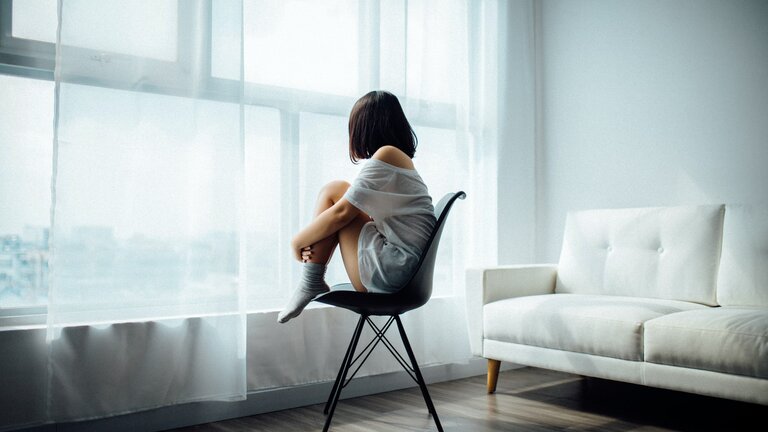 Eine Frau sitzt mit angezogenen Knien auf einem Stuhl und schaut aus dem Fenster. | © unsplash 