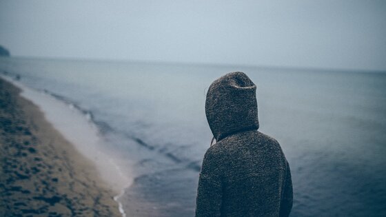 Eine Person mit einer Kaputze läuft alleine am Strand. | © Unsplash