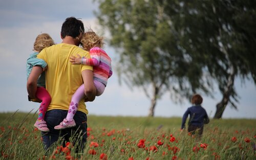 Ein Vater trägt seine zwei kleinen Kinder und läuft mit ihnen auf einem Blumenfeld.  | © unsplash