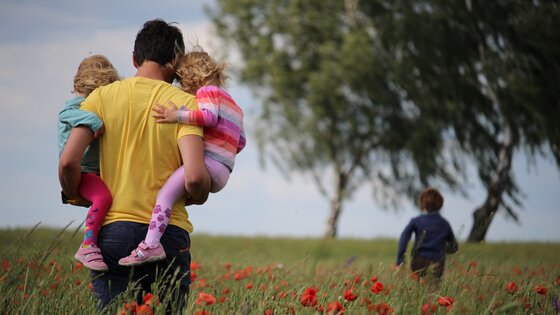 Ein Vater trägt seine zwei kleinen Kinder und läuft mit ihnen auf einem Blumenfeld.  | © Unsplash