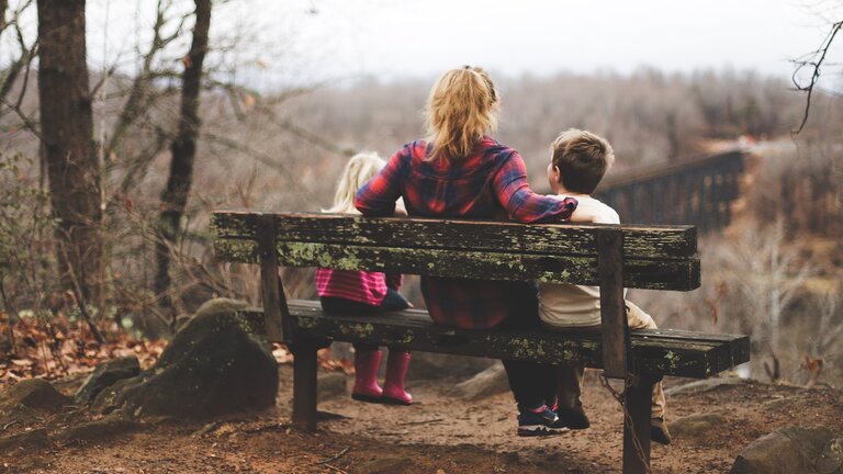 Eine Frau sitzt mit ihren zwei Kindern auf einer Bank in der Natur. | © Unsplash