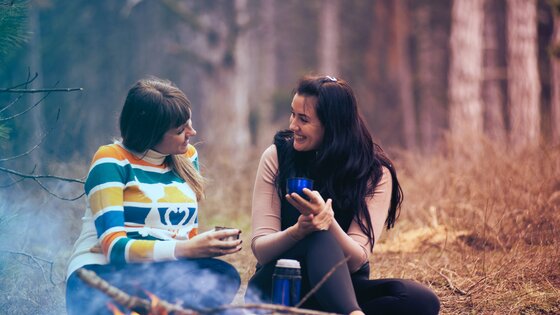 Zwei junge Frauen sitzen im Wald an einem Lagerfeuer und unterhalten sich miteinander. | © Pexels/ Alexandr Podvalny