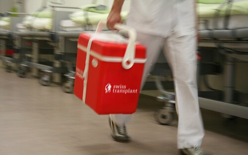 Auf dem Bild ist eine Transportbox von Swisstransplant zu sehen. Sie ist rot und mit dem weissen Logo versehen. Im Hintergrund ist ein Spital zu erkennen. | © Swisstransplant