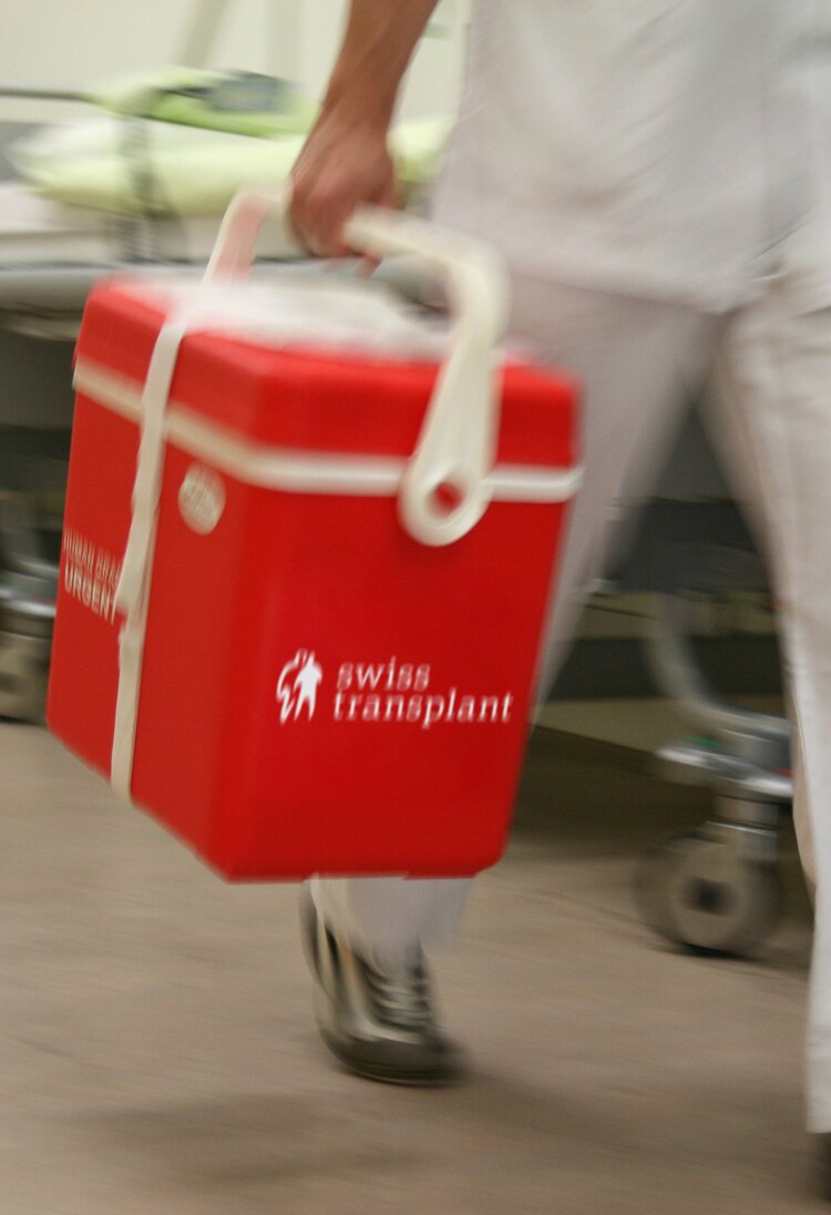 Auf dem Bild ist eine Transportbox von Swisstransplant zu sehen. Sie ist rot und mit dem weissen Logo versehen. Im Hintergrund ist ein Spital zu erkennen. | © Bild: Swisstransplant
