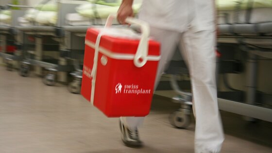 Auf dem Bild ist eine Transportbox von Swisstransplant zu sehen. Sie ist rot und mit dem weissen Logo versehen. Im Hintergrund ist ein Spital zu erkennen. | © Bild: Swisstransplant
