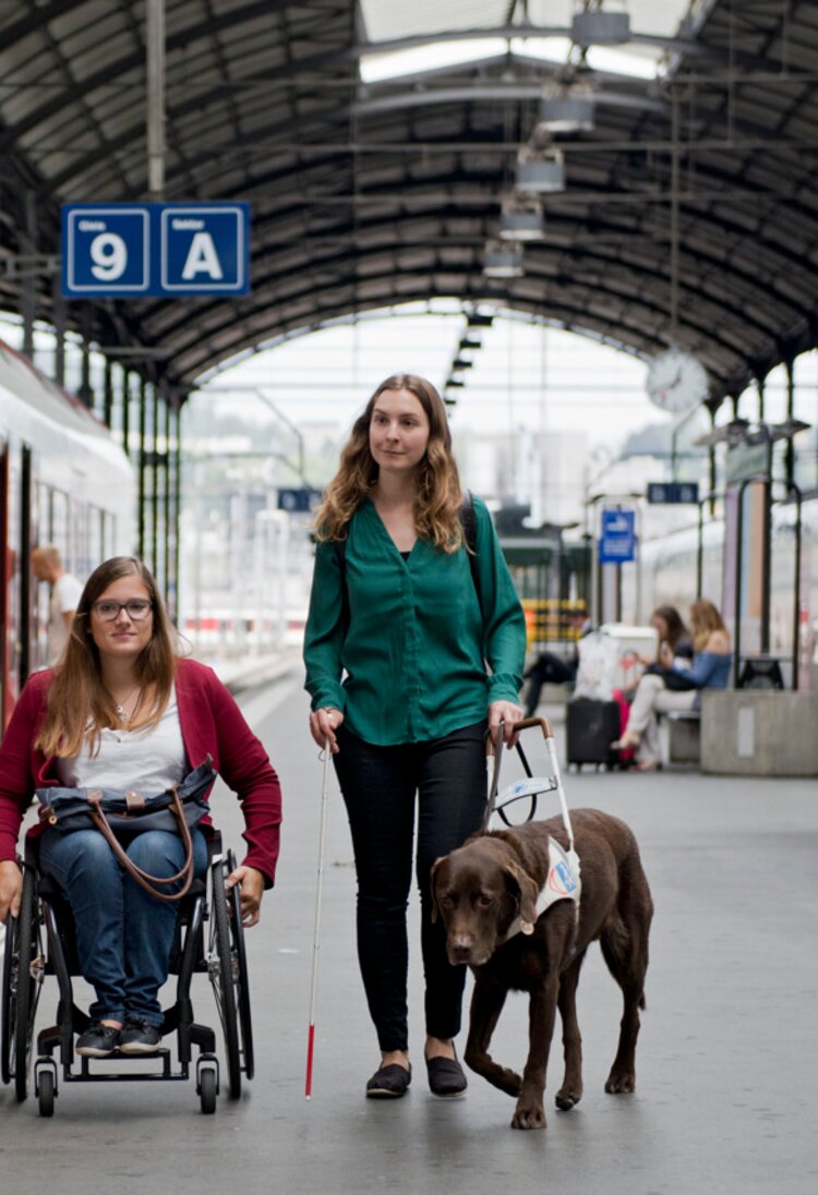 Eine Reisende im Handrollstuhl und eine blinde Reisende mit Blindenführhund auf dem Perron. | © SBB CFF FFS/Flavia Trachsel
