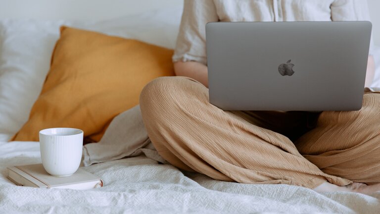 Eine Frau sitzt im Schneidersitz auf dem Bett, den Laptop auf den Beinen. Neben sich hat sie eine Tasse Kaffee. | © pexels